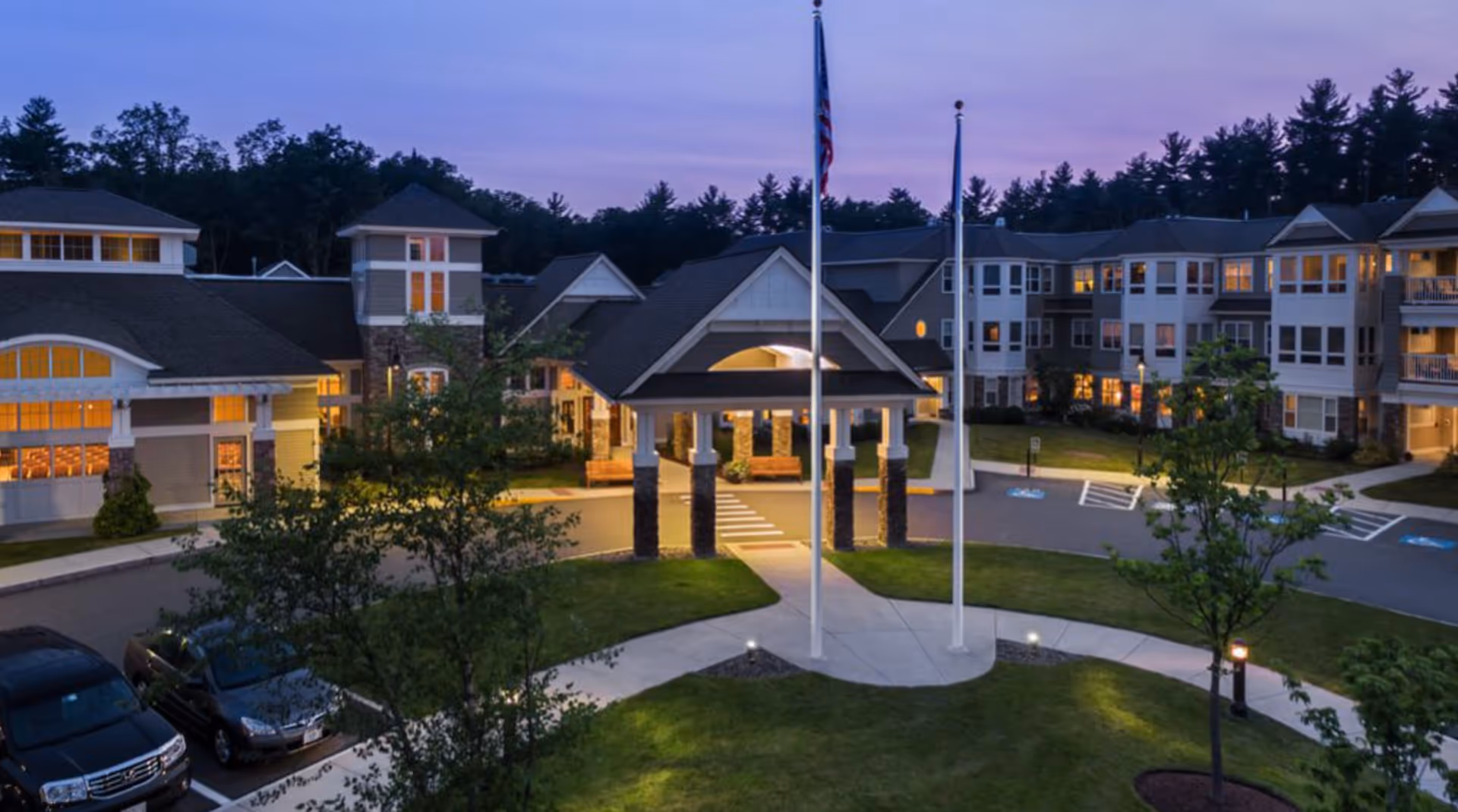 Front entrance and driveway of a multi-story senior living complex at dusk with flagpoles and illuminated windows.