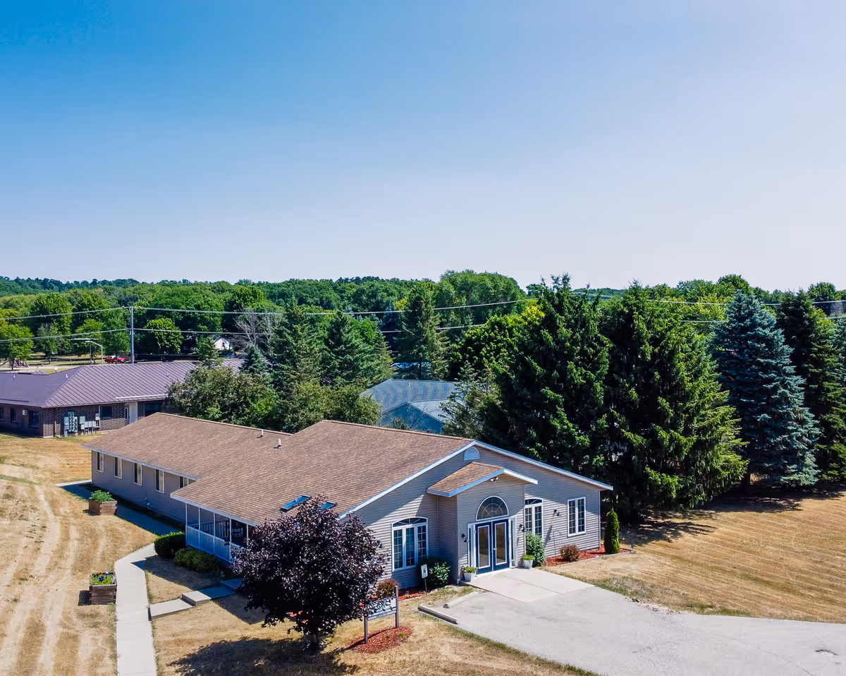 Aerial view of a single-story building with a brown roof and light-colored siding, surrounded by trees and a dry grassy area under a clear blue sky.