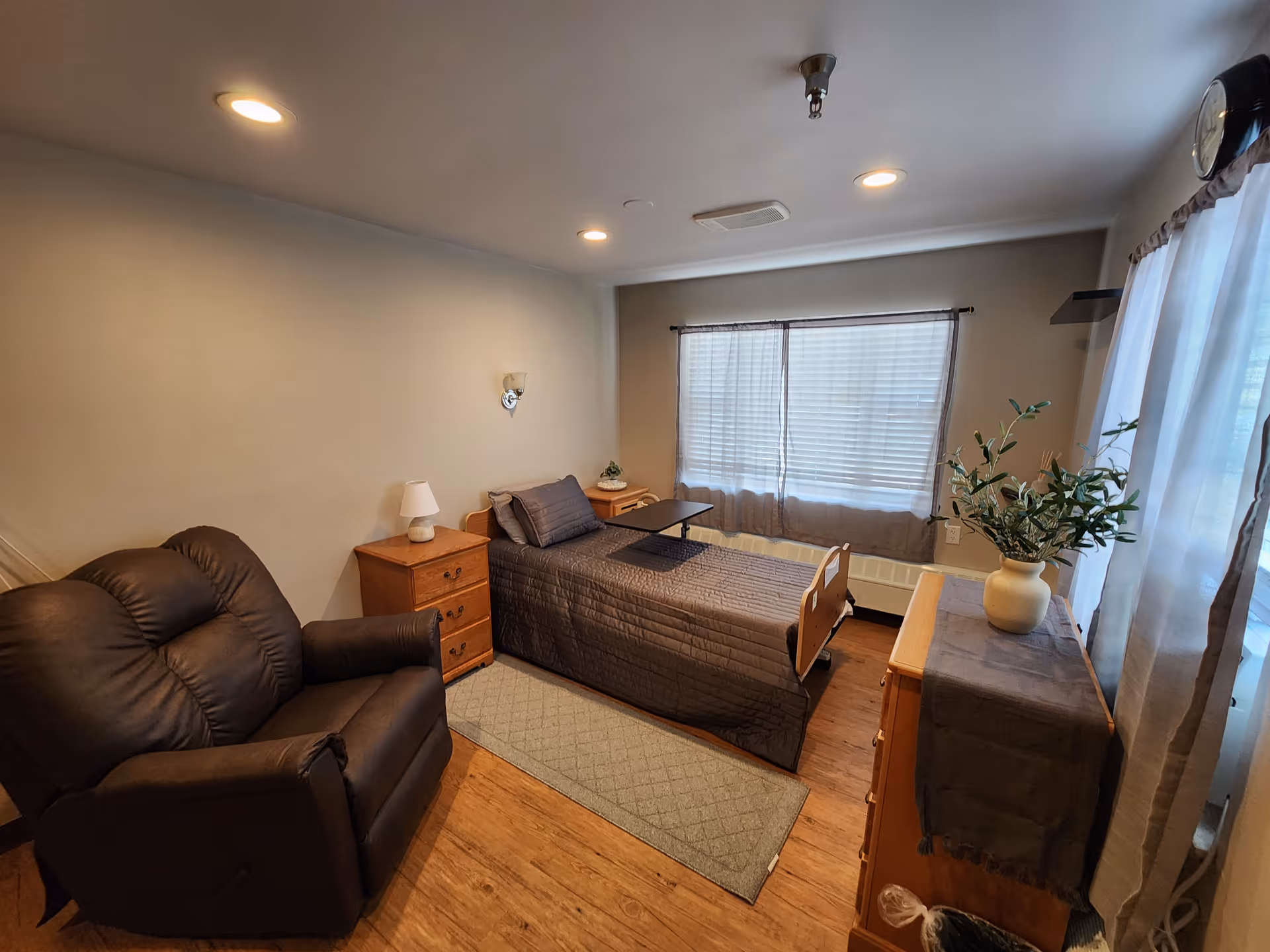 A single bed with nightstands, a brown recliner, dresser with plant, and a large window.