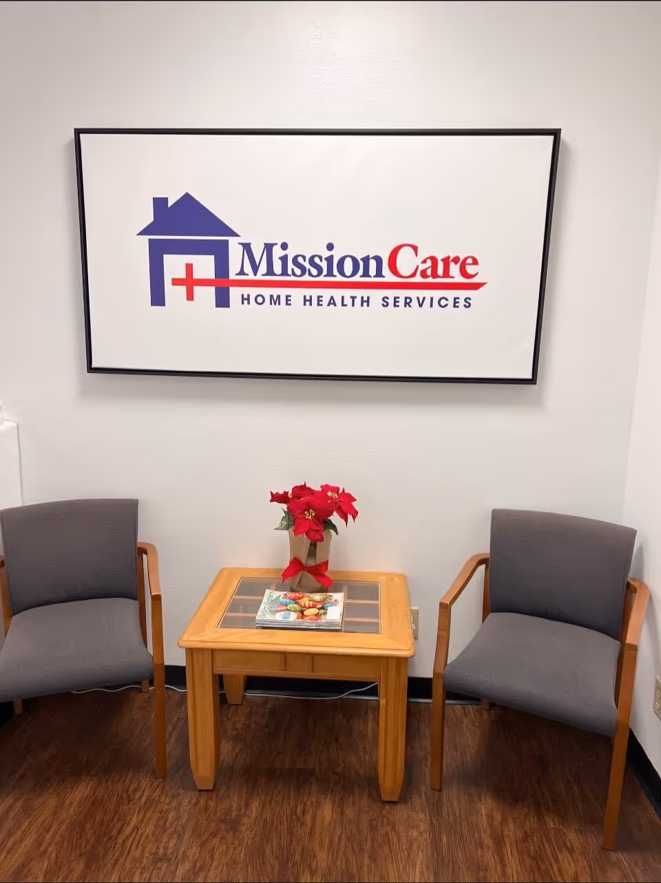 Interior lobby with two gray chairs, a wooden table, magazine, poinsettia, and MissionCare sign.