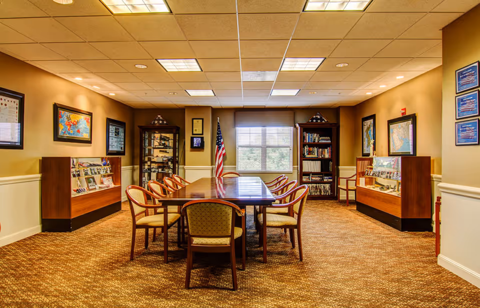 A well-lit meeting or conference room with a long wooden table surrounded by chairs. The room has beige walls with framed pictures and display cases along the sides. An American flag stands near a window with blinds, and there are bookshelves and display cabinets containing various items.