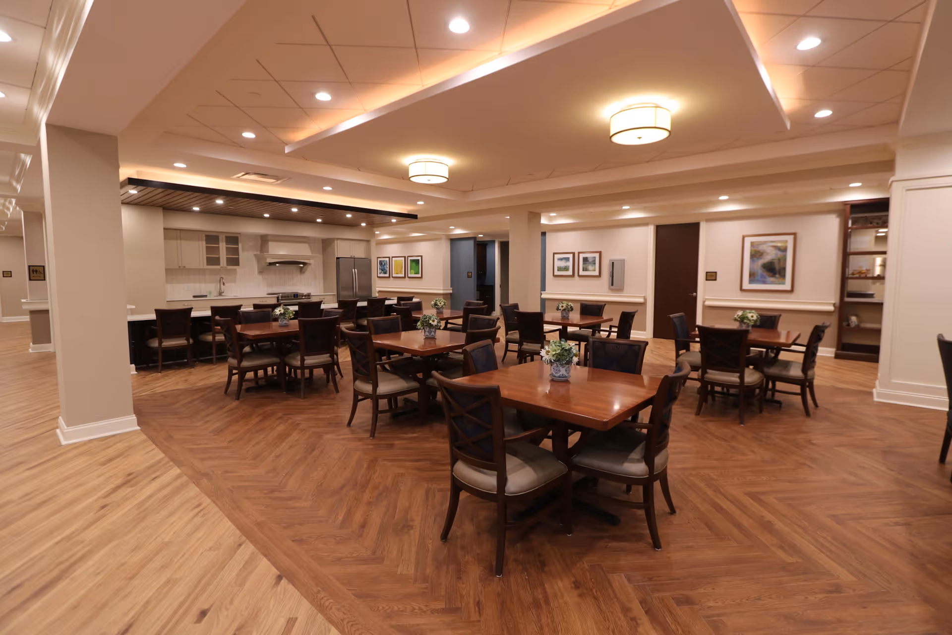 A spacious dining room with multiple wooden tables and chairs arranged neatly. Each table has a small floral centerpiece. The room features a modern kitchen area in the background with a refrigerator, stove, and cabinetry. The floor has a wood pattern, and the ceiling has recessed lighting and two round ceiling lights. The walls are decorated with framed artwork.