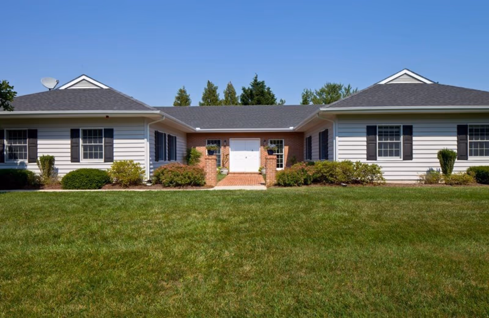 Single-story white-clad building with a central entrance, symmetrical wings, shrubs, and a large front lawn under a clear blue sky.