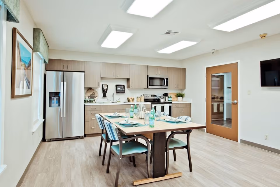 A bright and modern kitchen and dining area with light wood flooring, light wood cabinets, a stainless steel refrigerator, microwave, and stove. A wooden dining table with four chairs is set with plates, glasses, and bottled water. The room has white walls, a wall-mounted TV, and a door with a window leading to another room.