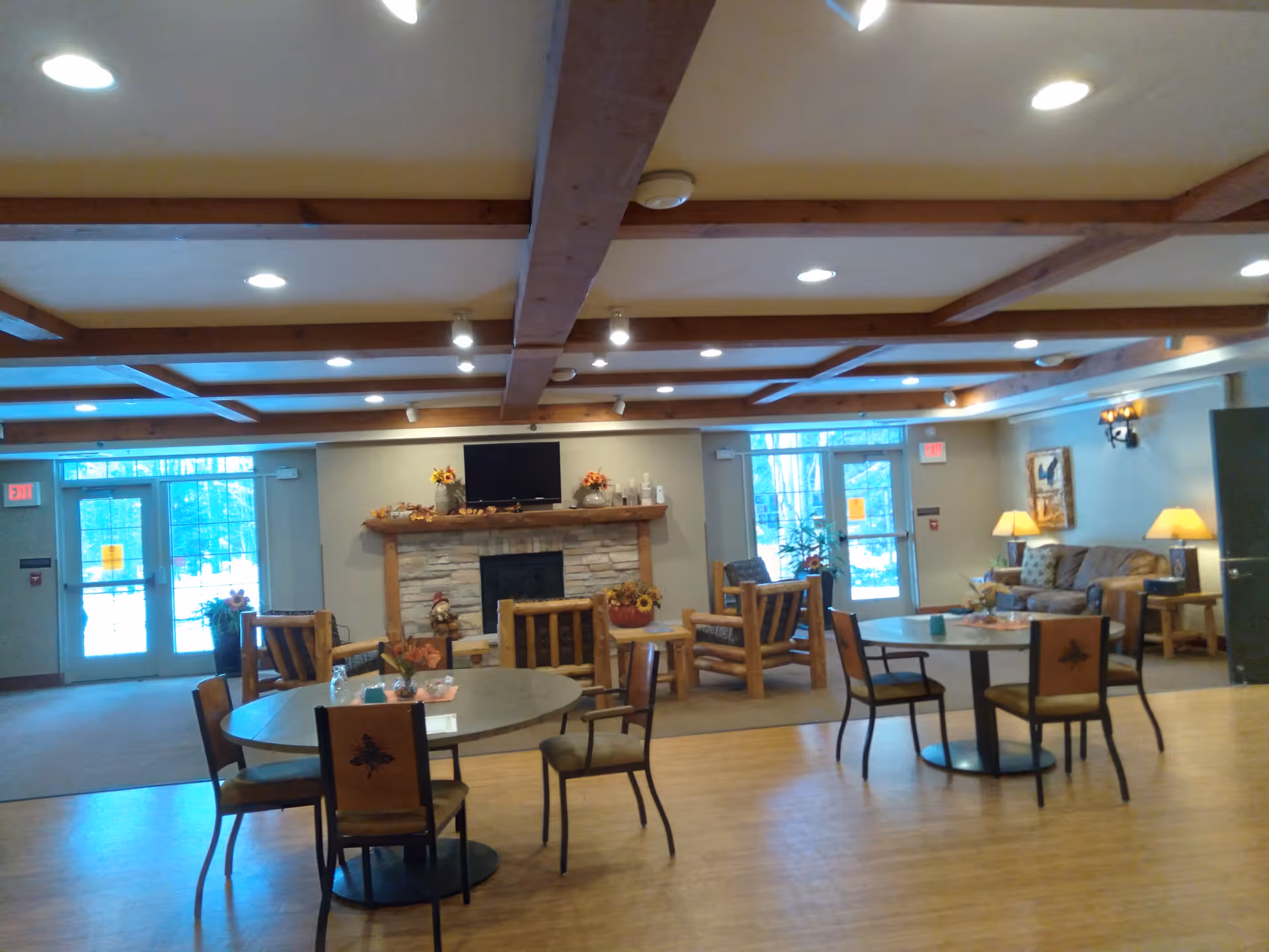 Spacious common room with round dining tables and chairs, a seating area with sofas and a fireplace under exposed wooden ceiling beams.
