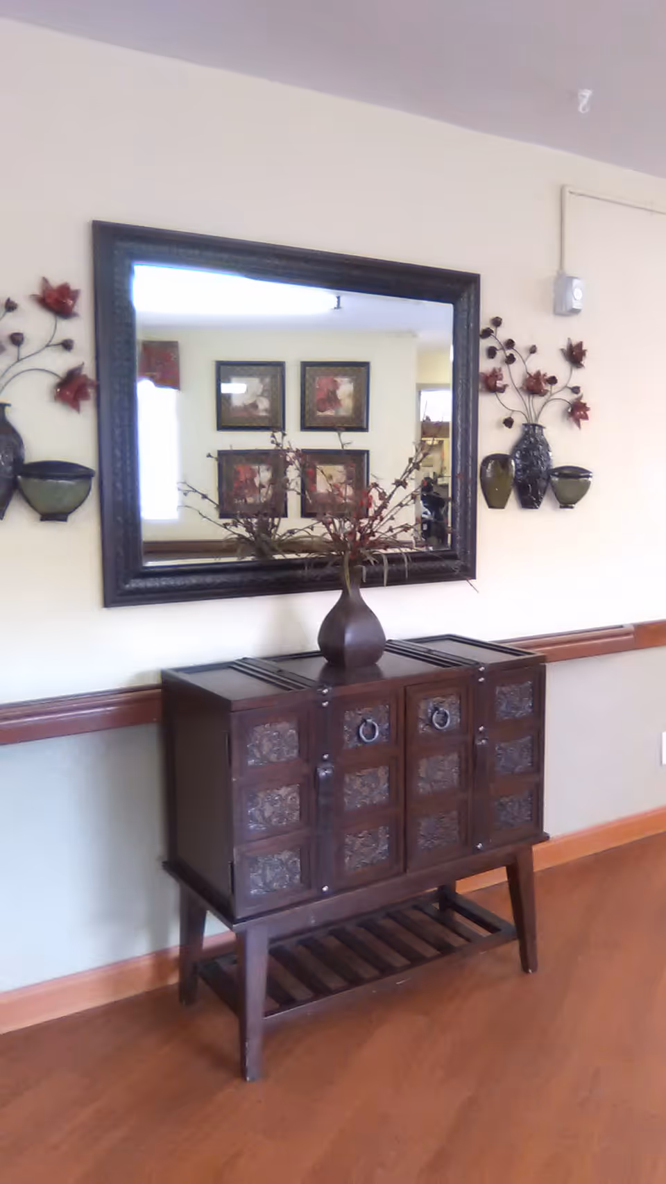 Dark wooden console cabinet with decorative mirror and metal wall art along a hallway.