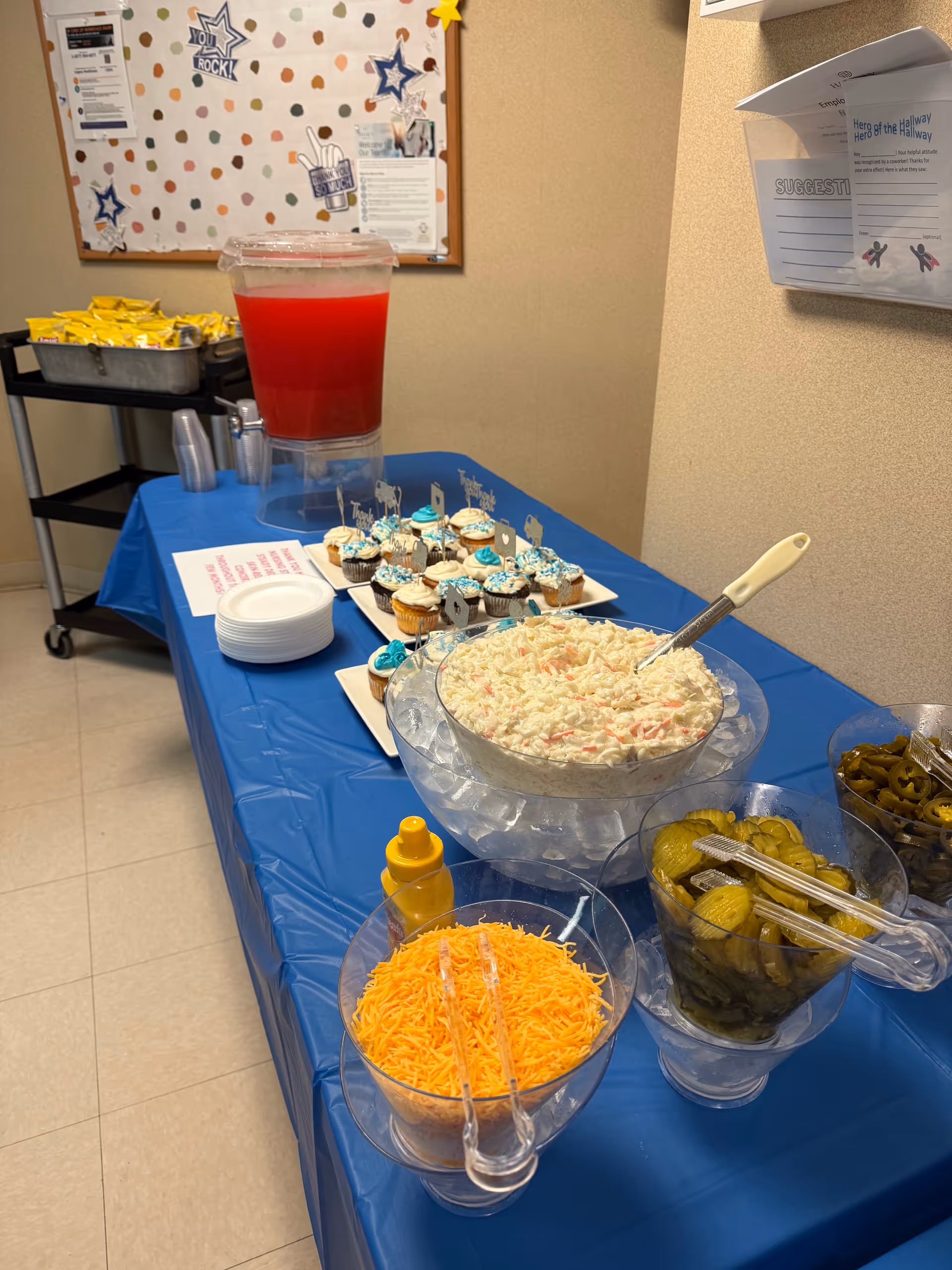 Buffet table in an indoor common area with bowls of coleslaw, shredded cheese, pickles, cupcakes and a red drink dispenser.