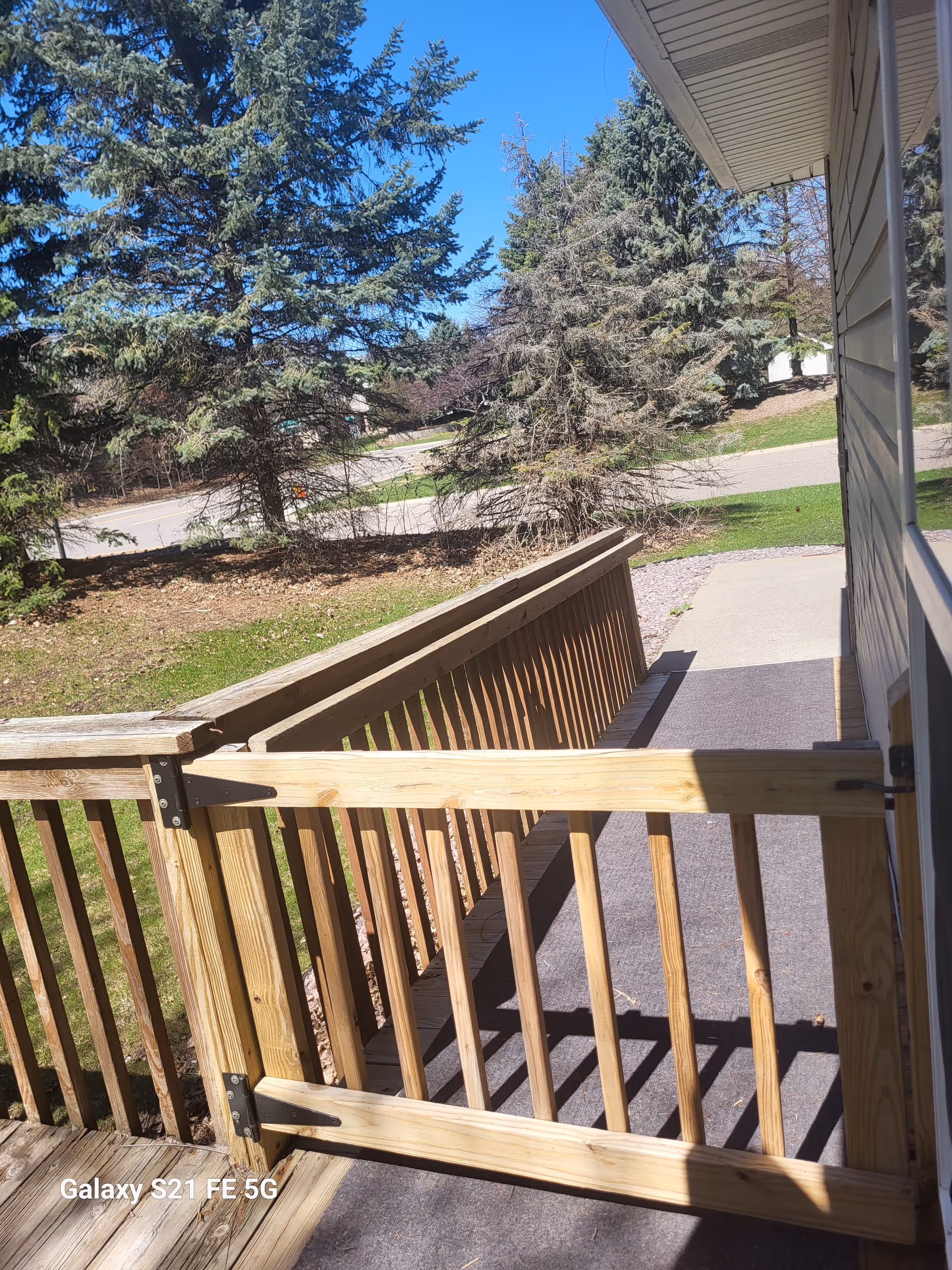 Wooden deck railing along a building with a paved walkway and evergreen trees nearby