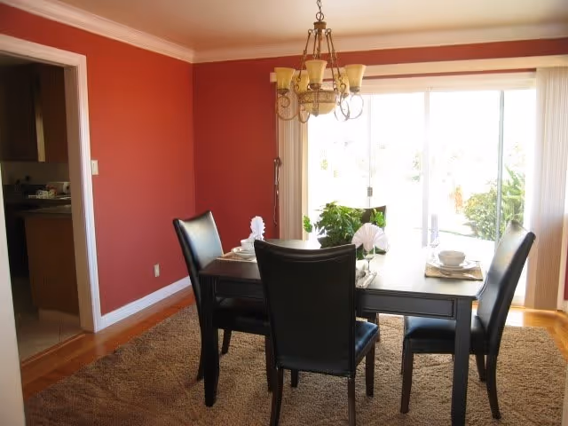 Dining room with a dark wooden table set for four with black leather chairs, a chandelier hanging above, red walls, a large window with vertical blinds letting in natural light, and a view of greenery outside.