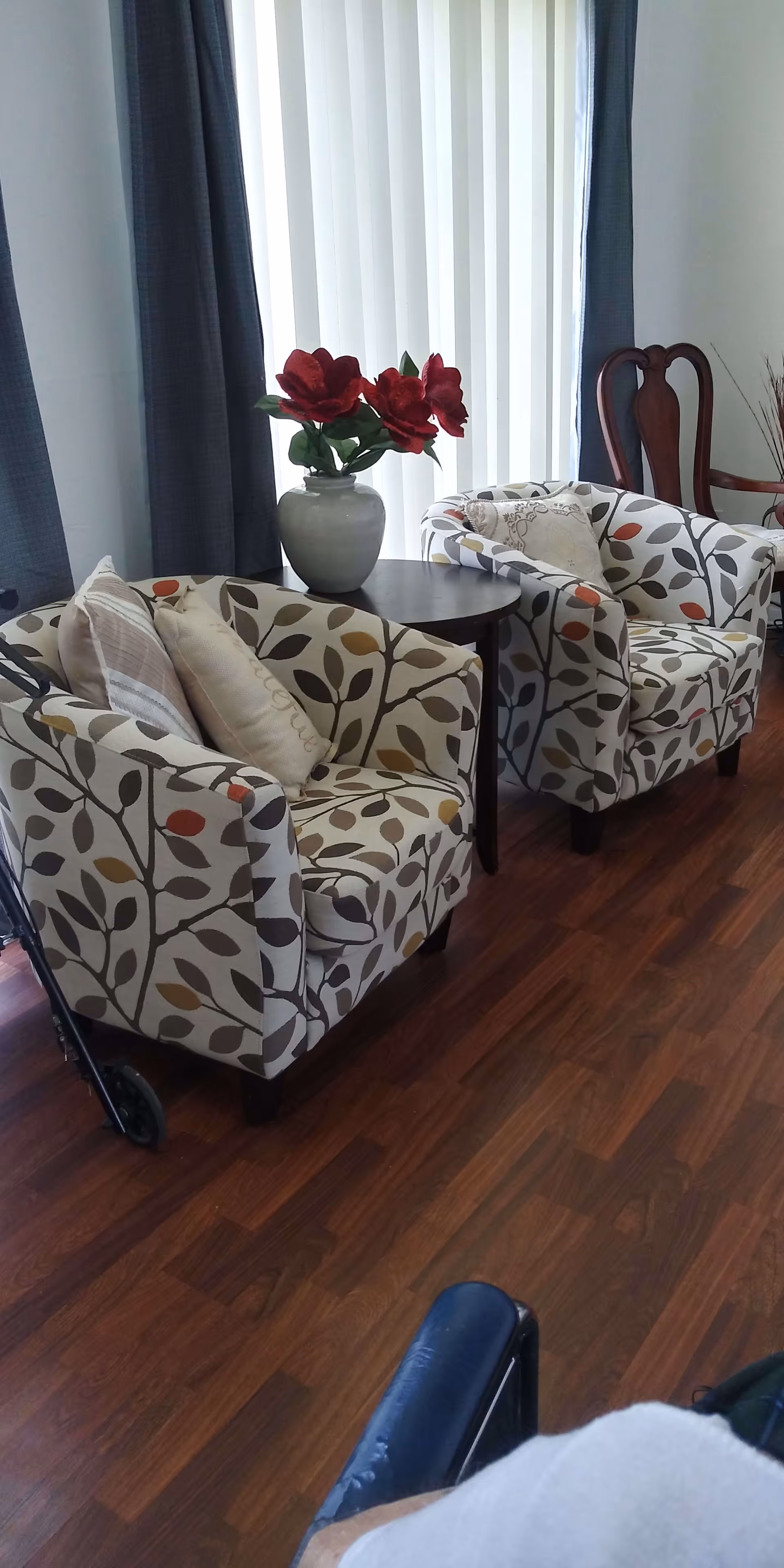 Two patterned armchairs with leaf designs and cushions are placed around a small round wooden table with a vase of red flowers on it. Behind the chairs are tall windows with vertical blinds and dark curtains. A wooden chair with a carved backrest is visible in the background, and part of a wheelchair is seen in the foreground on a wooden floor.