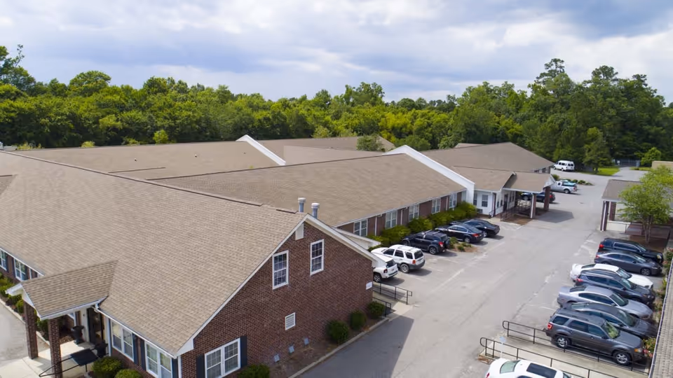 Aerial view of Compass Post Acute Rehabilitation facility showing a large single-story brick building with a beige shingled roof, surrounded by a parking lot with several parked cars and trees in the background under a partly cloudy sky.