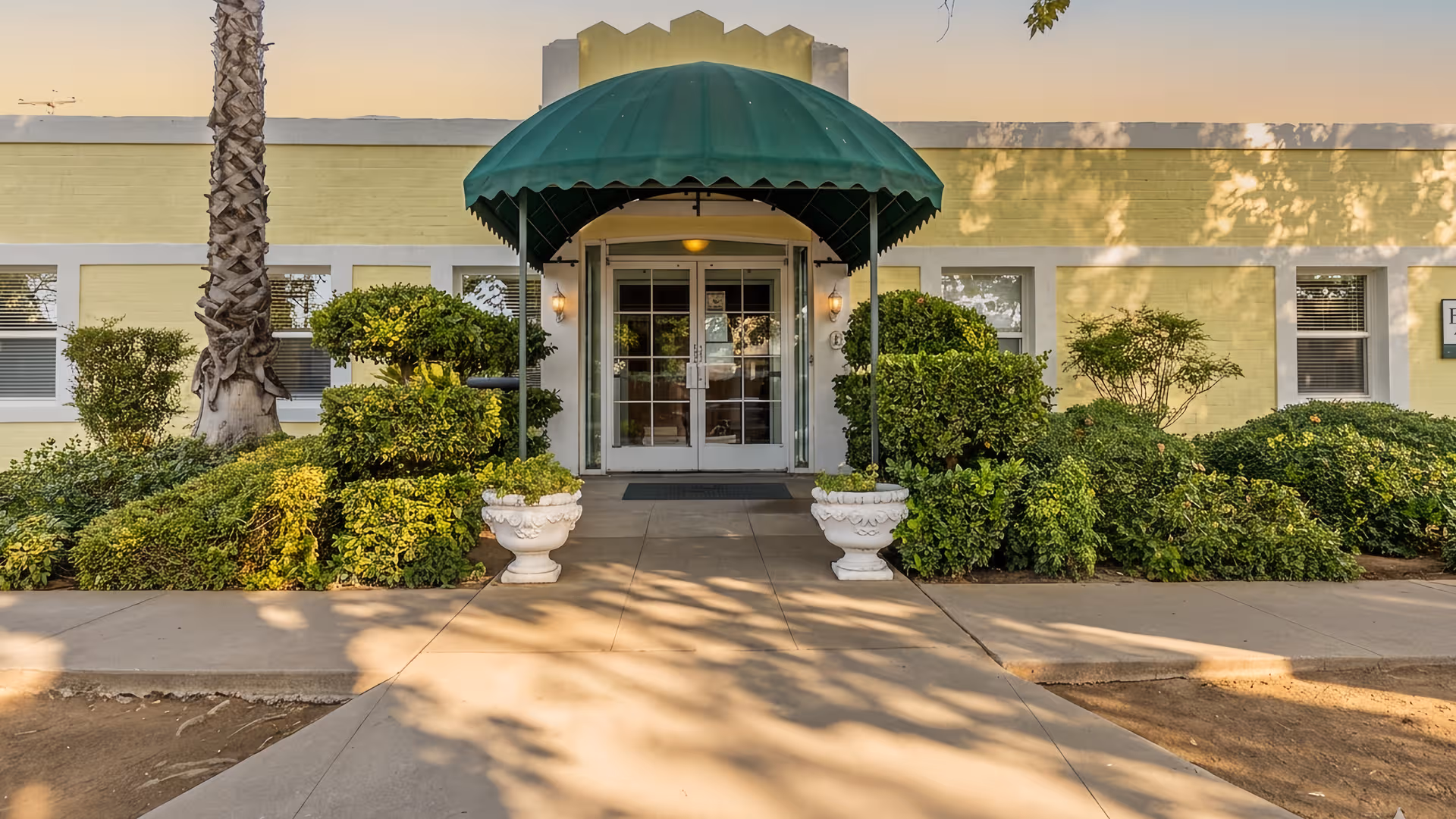 Front entrance of a senior living facility with a green canopy and manicured hedges.