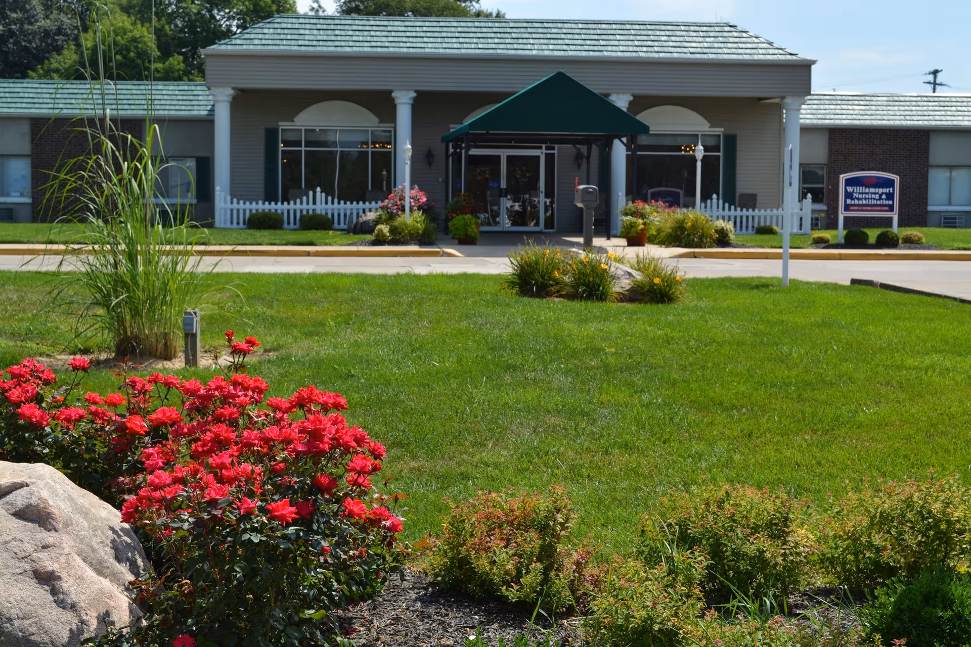 Front entrance of a nursing and rehabilitation facility with a covered doorway, green lawn and flower beds in the foreground.