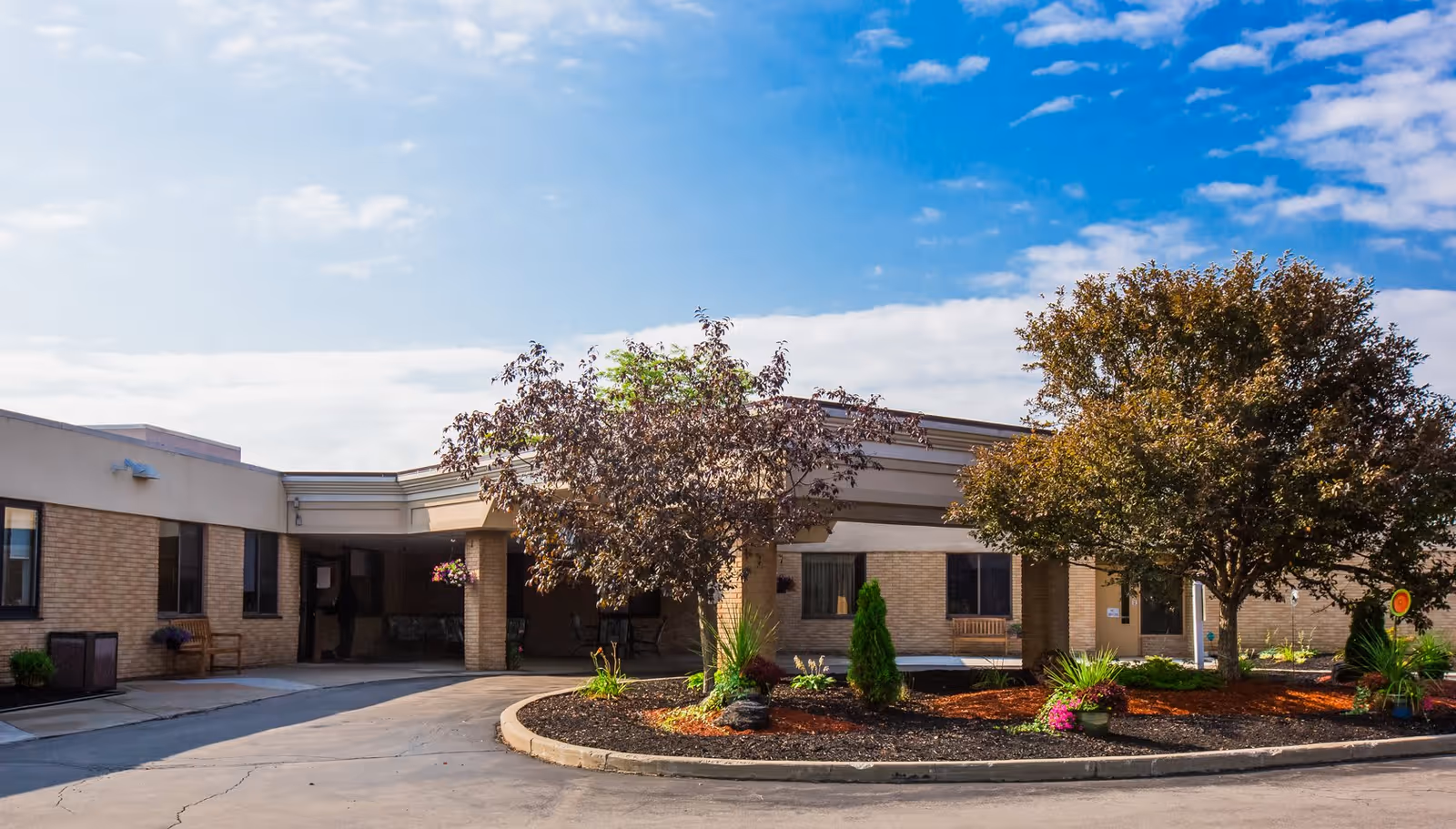 Front entrance of a single-story senior living facility with a circular driveway, landscaped island and trees under a blue sky.