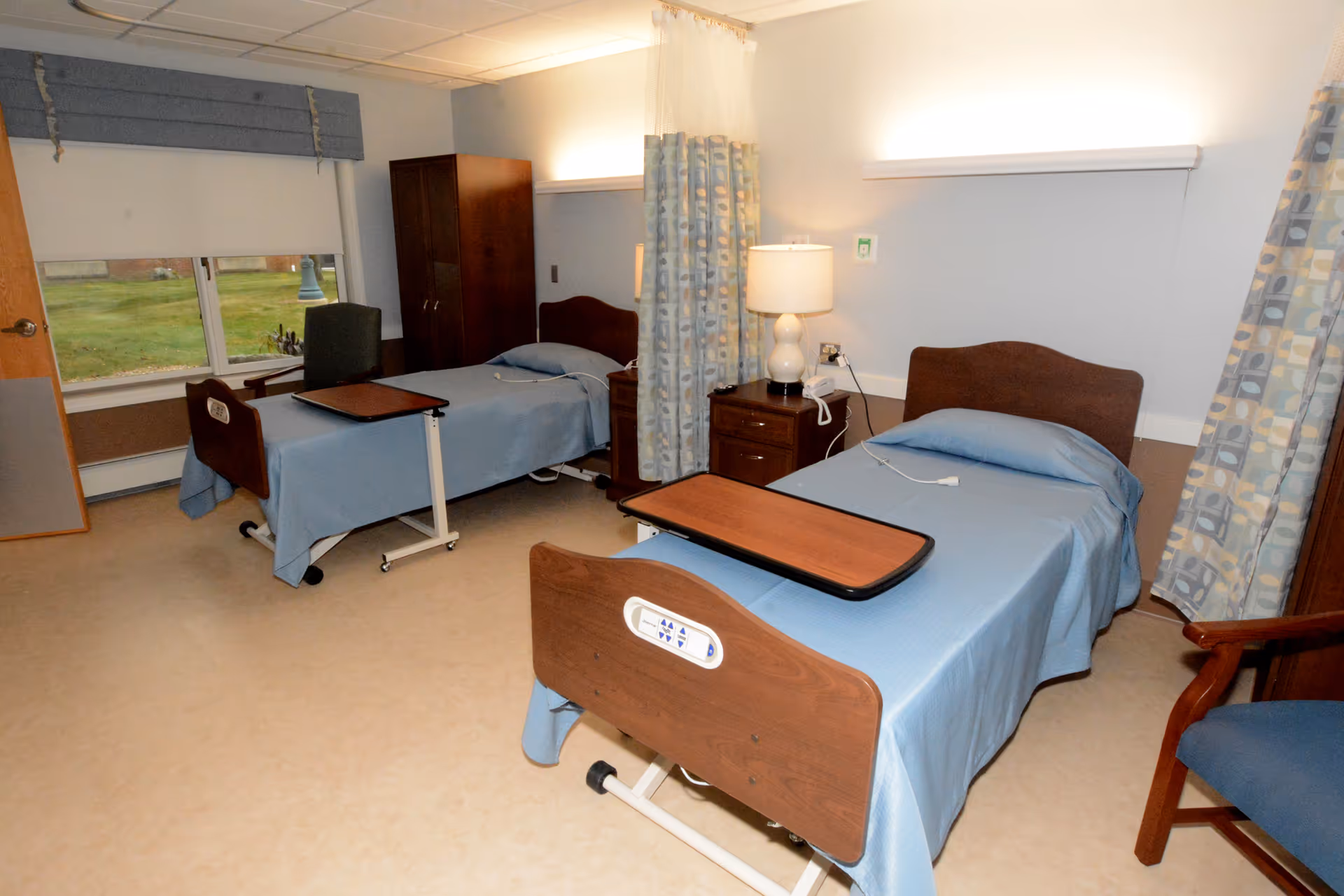A shared patient room in a healthcare facility with two hospital beds covered in blue bedding, each equipped with an overbed table. The room has a nightstand with a lamp and a telephone between the beds, a wooden wardrobe, a chair, and windows with patterned curtains and a view of the outside lawn.