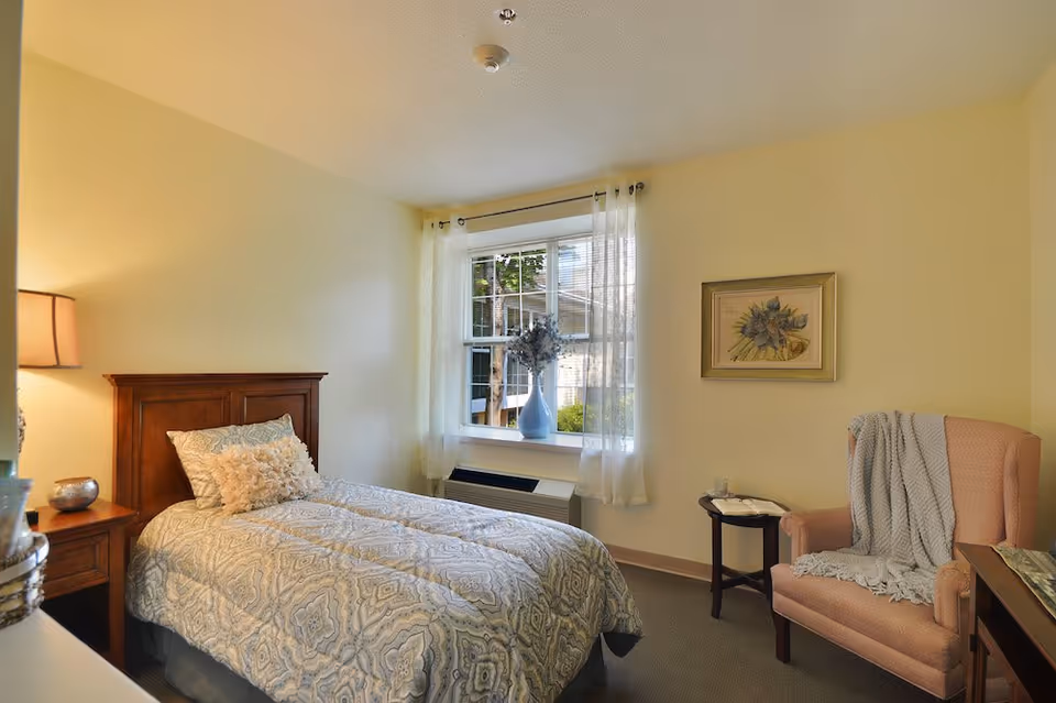 Cozy bedroom in a senior living facility with a bed, chair, window, and artwork.