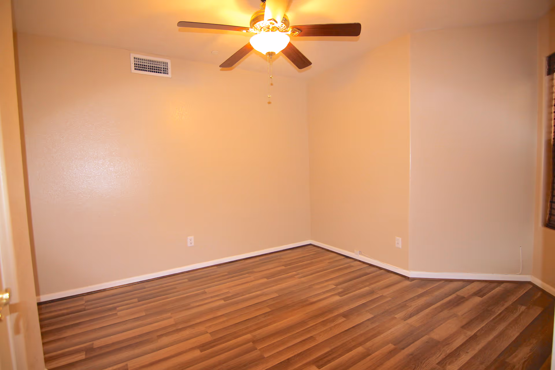 Empty room with beige walls, wood-patterned flooring, a ceiling fan with light, and a window with blinds partially visible on the right side.