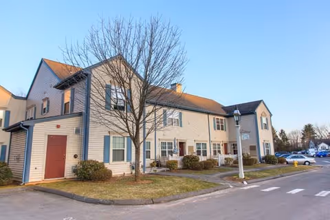 Exterior view of a two-story senior living facility building with beige siding and blue shutters. There is a leafless tree in front, a lamp post near the sidewalk, and several parked cars in the background under a clear blue sky.