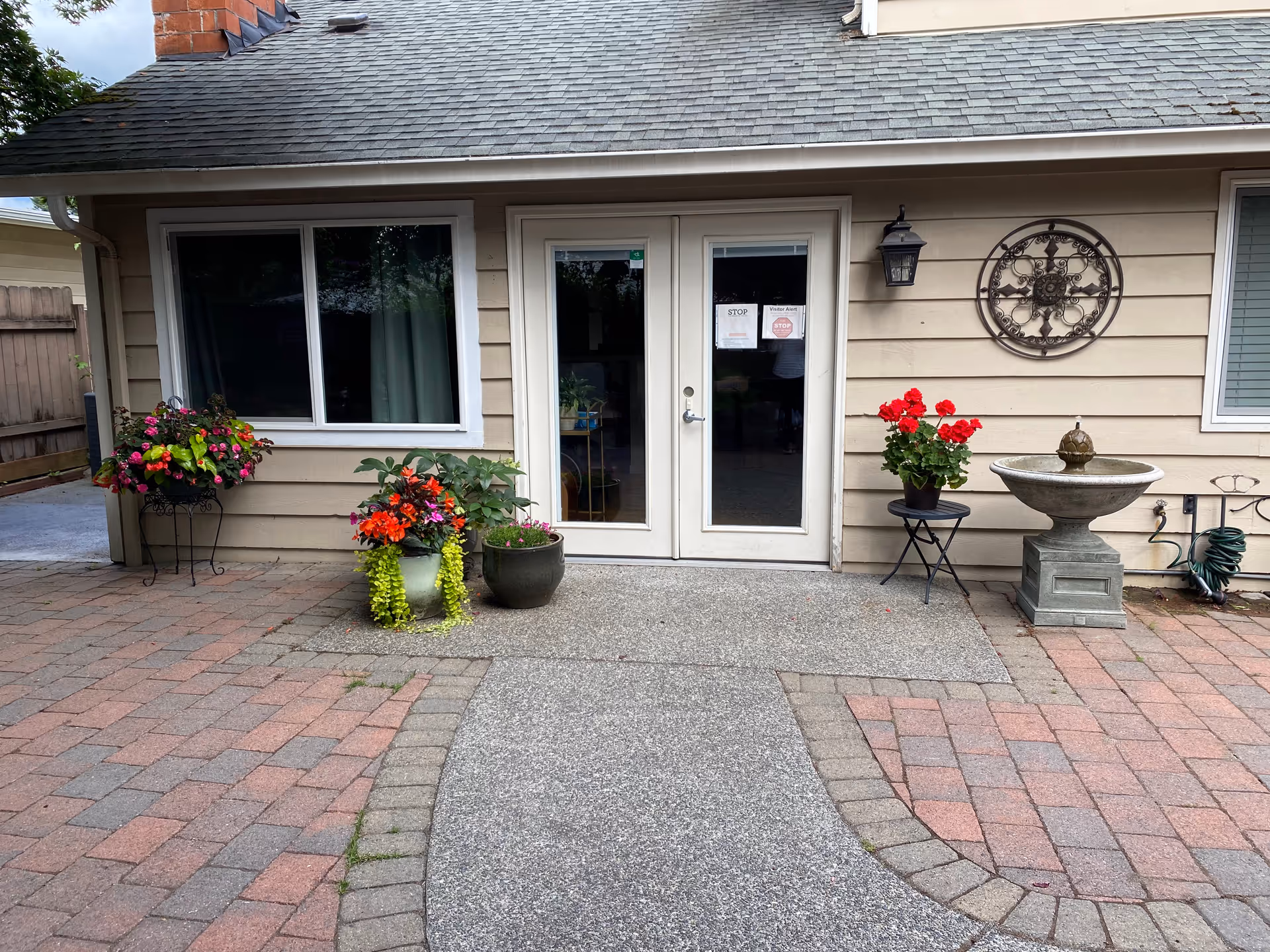 Exterior view of a senior living facility entrance with double glass doors, beige siding, a window on each side, decorative wall art, a wall-mounted lantern, a stone water fountain, and several potted plants with colorful flowers arranged on a brick and concrete paved area.