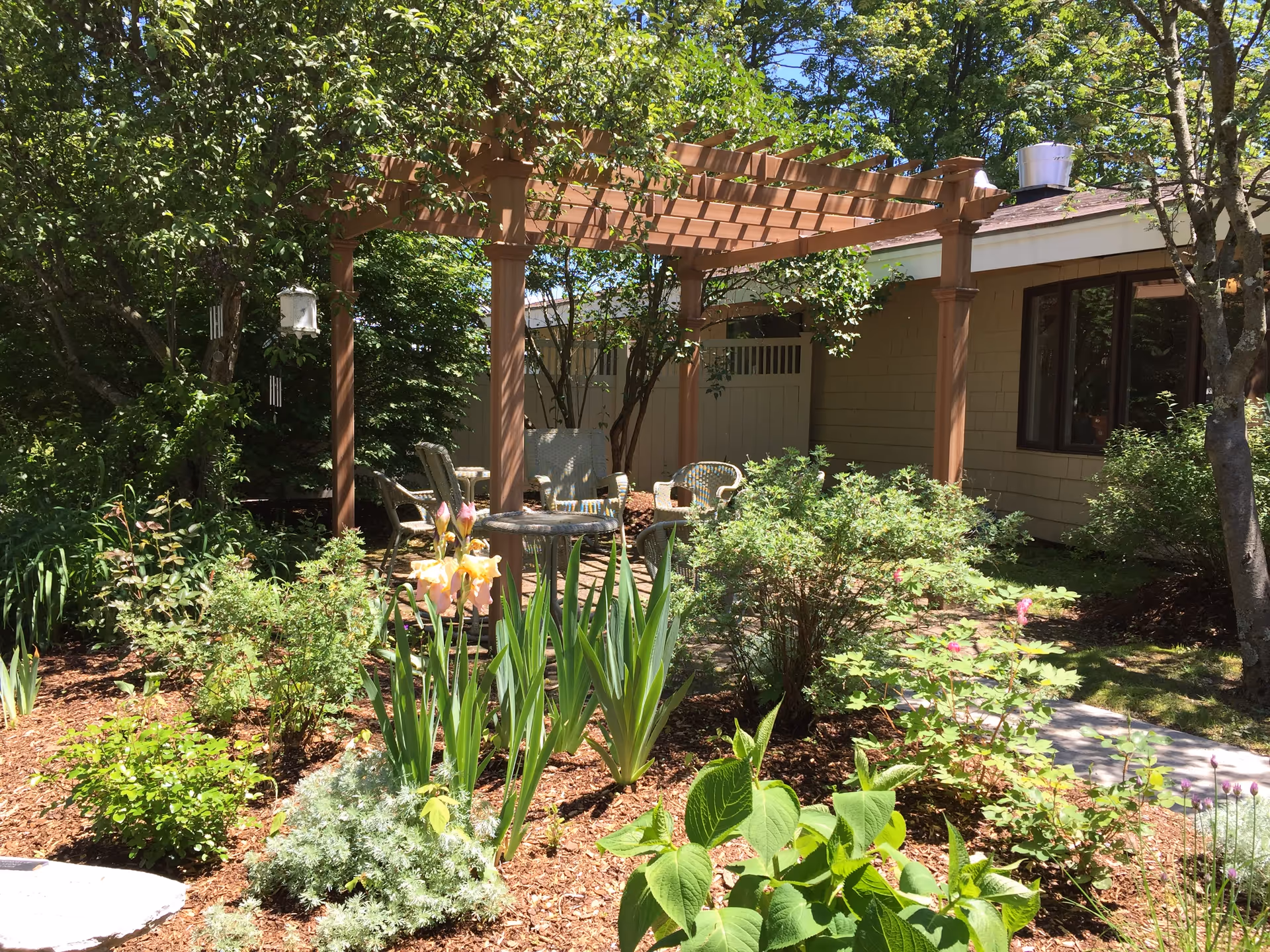 A sunny outdoor garden area with a wooden pergola providing partial shade over a seating arrangement of wicker chairs and a small table. The garden is lush with various green plants, shrubs, and blooming flowers. A building with beige siding and windows is visible in the background.