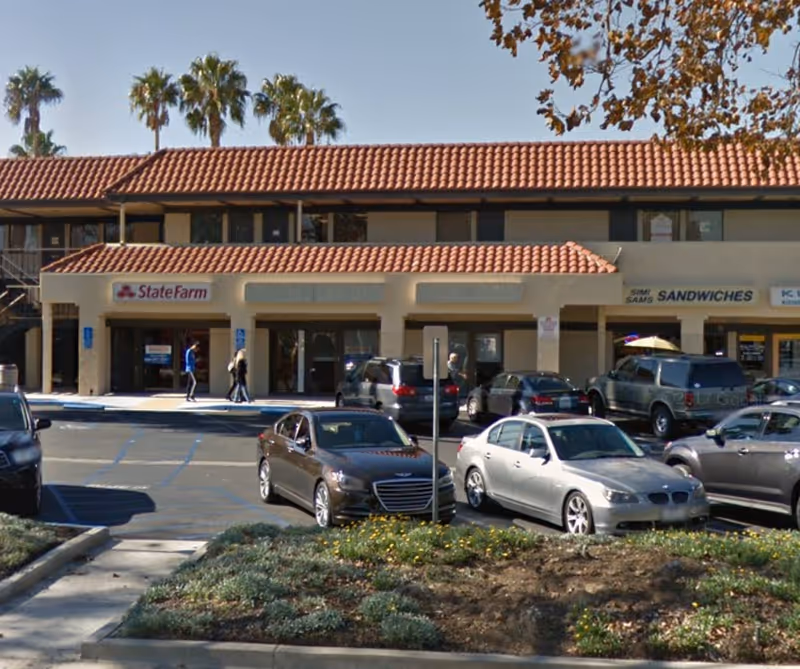 Exterior of a strip mall with beige storefronts, parked cars, and palm trees