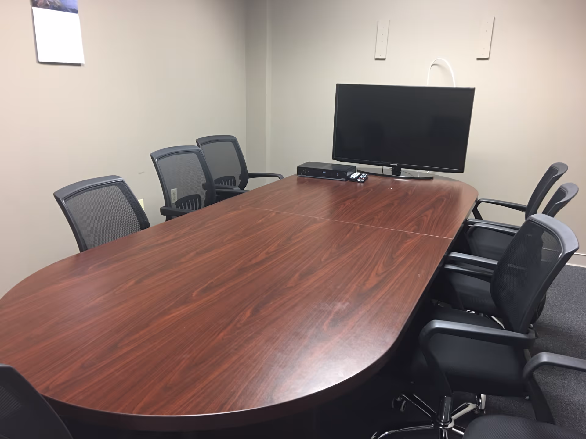 Empty boardroom with an oval wooden table, chairs, and a wall-mounted monitor.