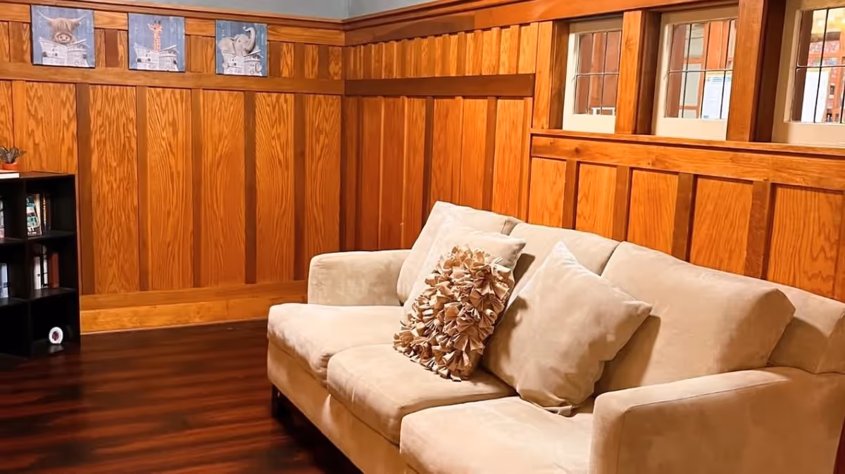 A cozy living room area with a beige sofa adorned with two decorative pillows, set against wood-paneled walls with three small windows above. There is a small black bookshelf with books and decorative items on the left side, and three framed animal pictures hanging on the wall above the sofa.