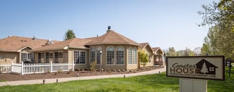 Exterior view of Conifer House, a single-story residential care and memory care facility with beige siding, multiple windows, a white picket fence, and a well-maintained lawn under a clear blue sky.
