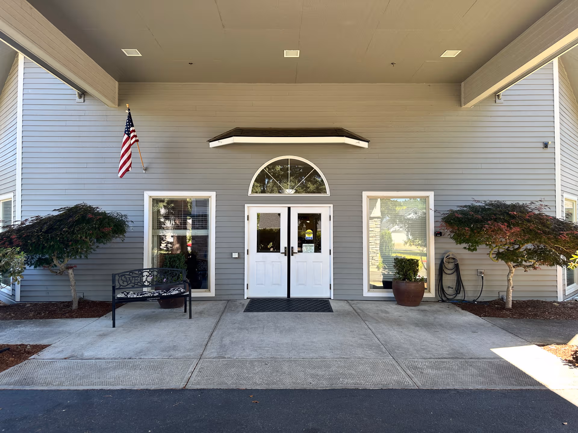 Front entrance to a senior living facility with double doors, flag, windows, and plants.