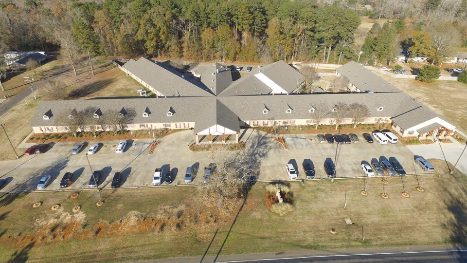 Aerial view of Arbor Lake Skilled Nursing and Rehabilitation Center showing a large, single-story building with a cross-shaped layout surrounded by parking spaces with several cars parked. The building is situated in a grassy area with trees in the background.
