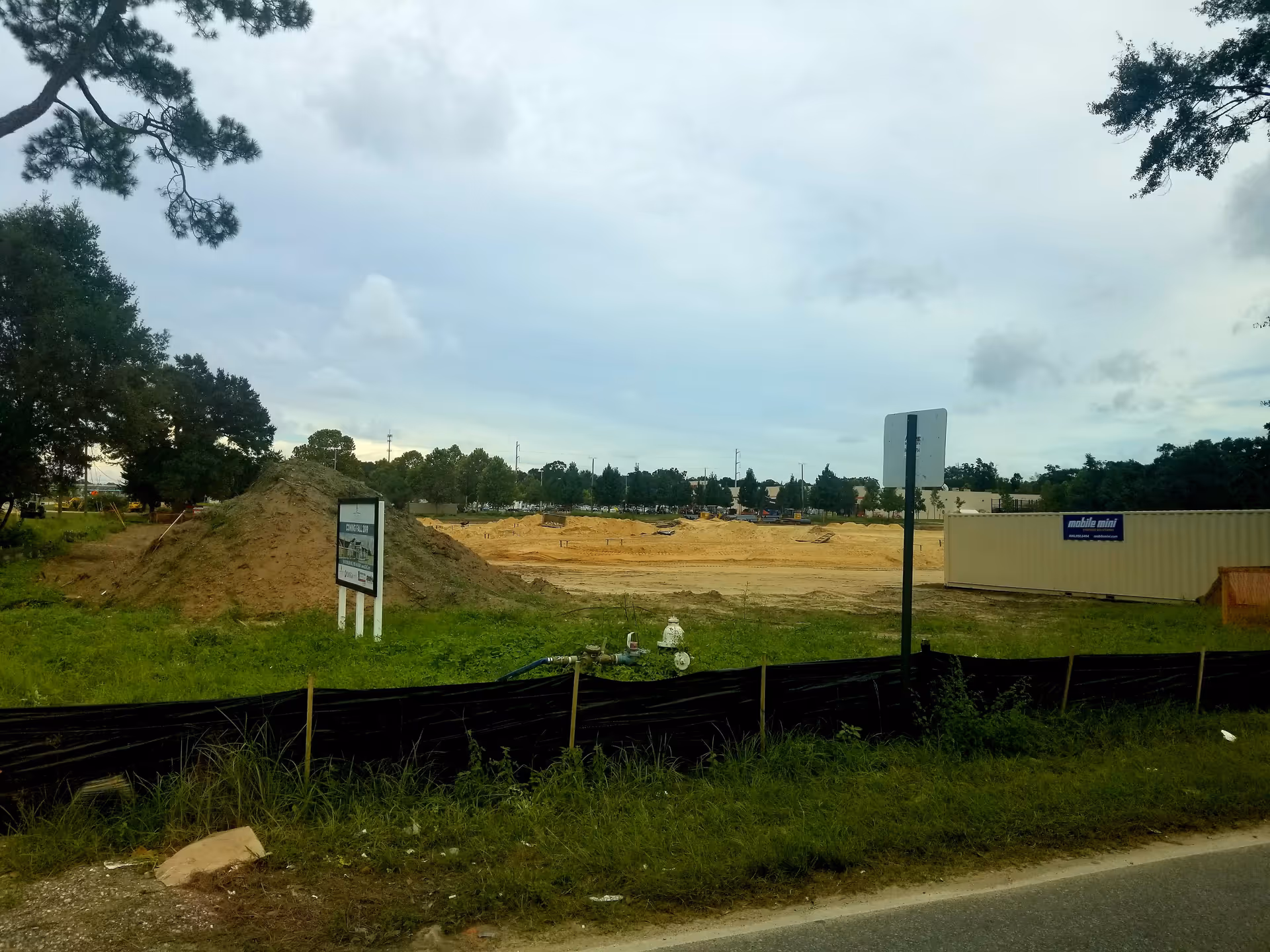 Construction site with dirt piles, fencing, and a portable building along the road.
