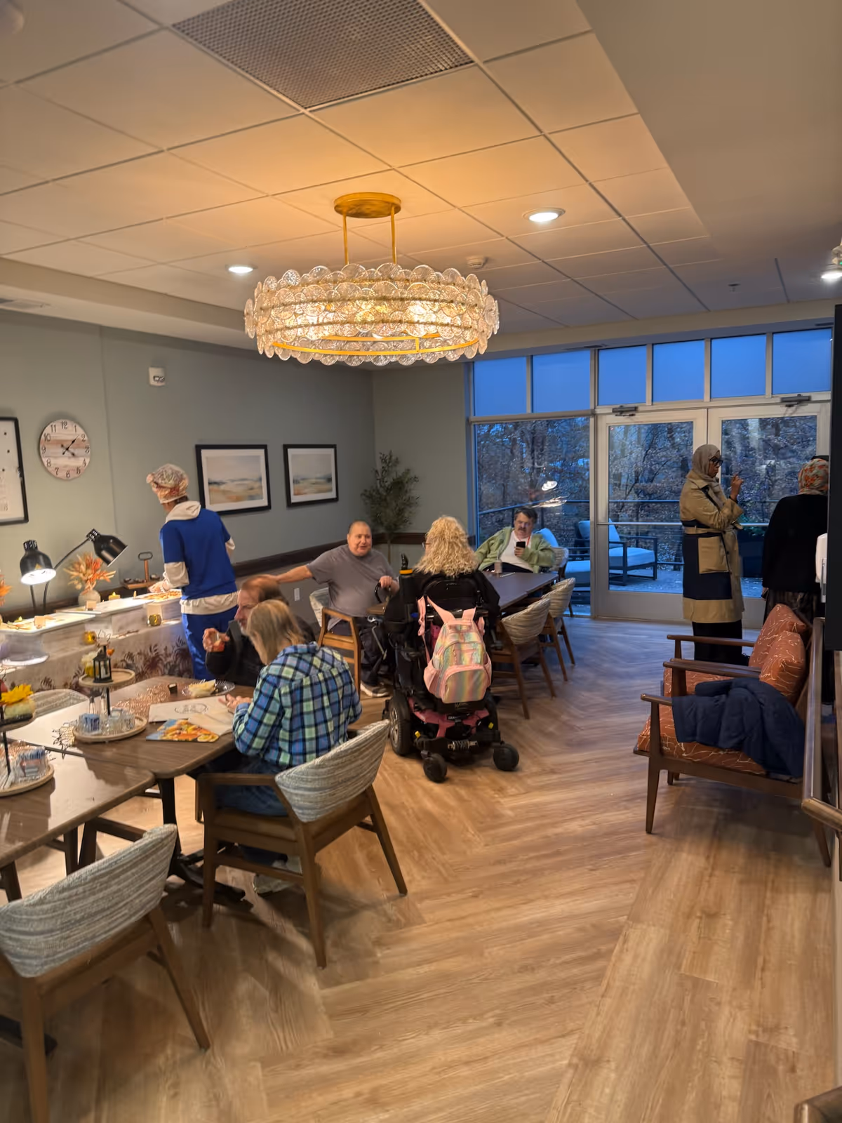 Residents seated at tables in a well-lit dining room with a chandelier and buffet.