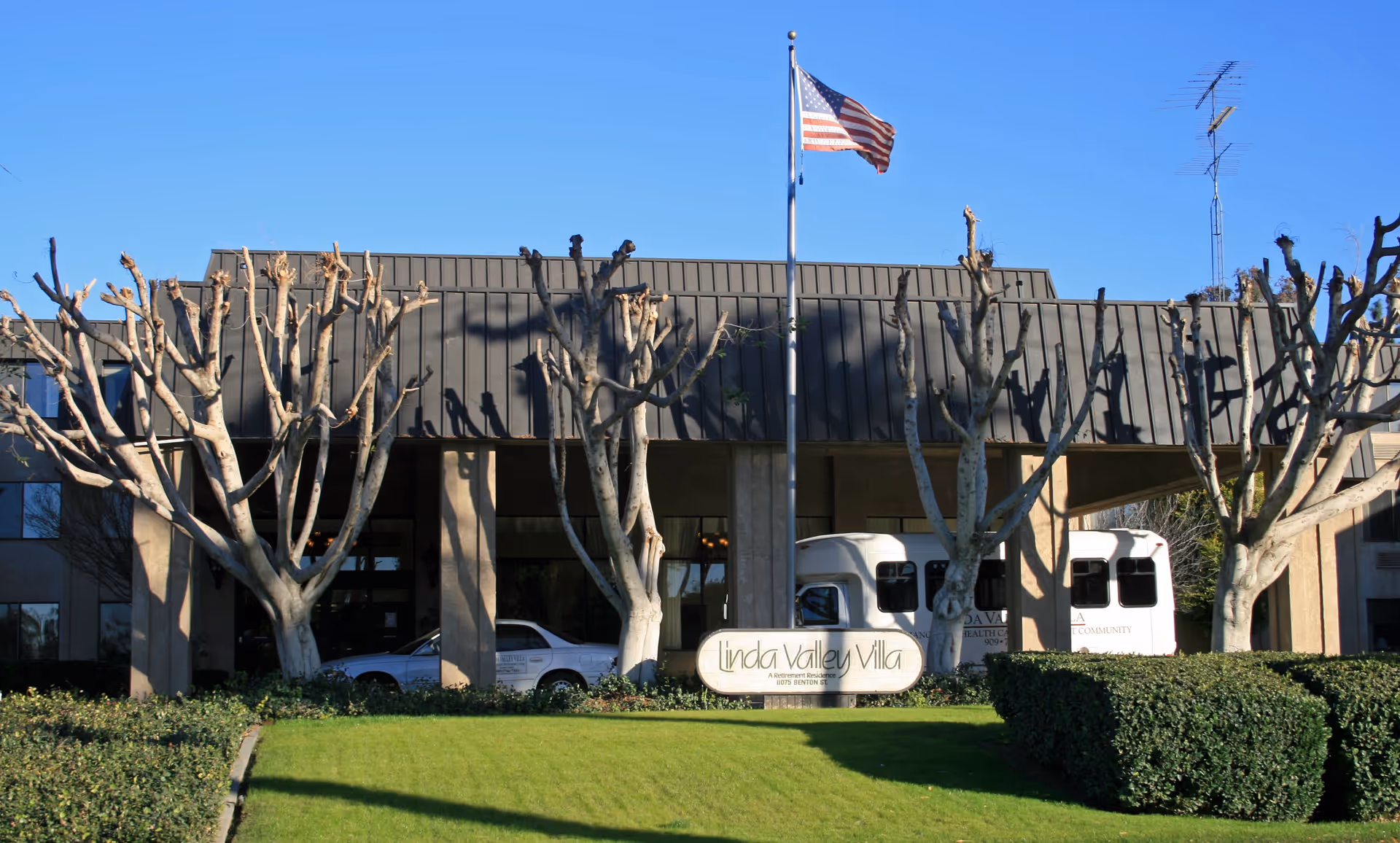 Front exterior of the Linda Valley Villa building with an American flag, pruned trees, and vehicles by the entrance.