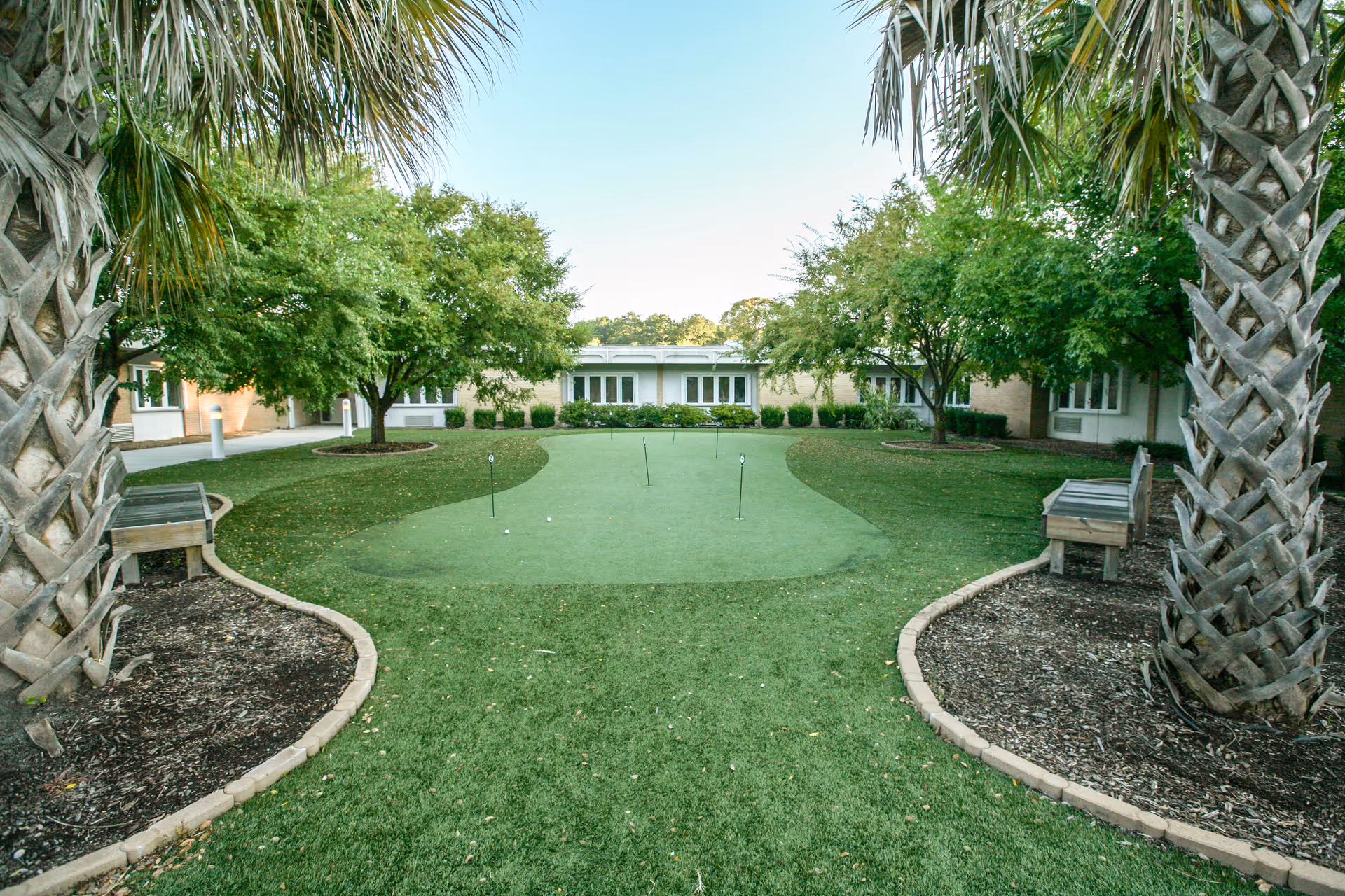 Outdoor putting green surrounded by grass, trees, and benches with a building in the background under a clear sky.