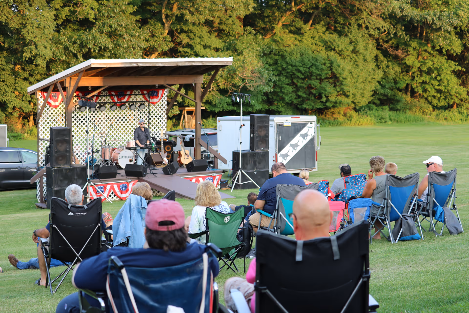 Outdoor lawn concert with audience seated in folding chairs watching a stage performance.