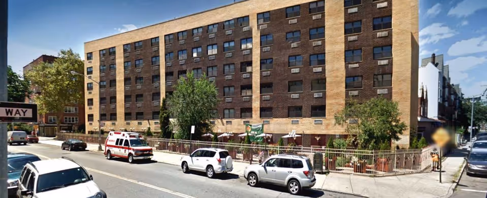 A multi-story brick senior living facility on a city street with parked cars.