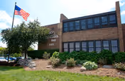 Exterior view of Plymouth Terrace, a two-story brick building with large windows. There is a landscaped area with bushes and a tree in front, and an American flag flying on a flagpole to the left.