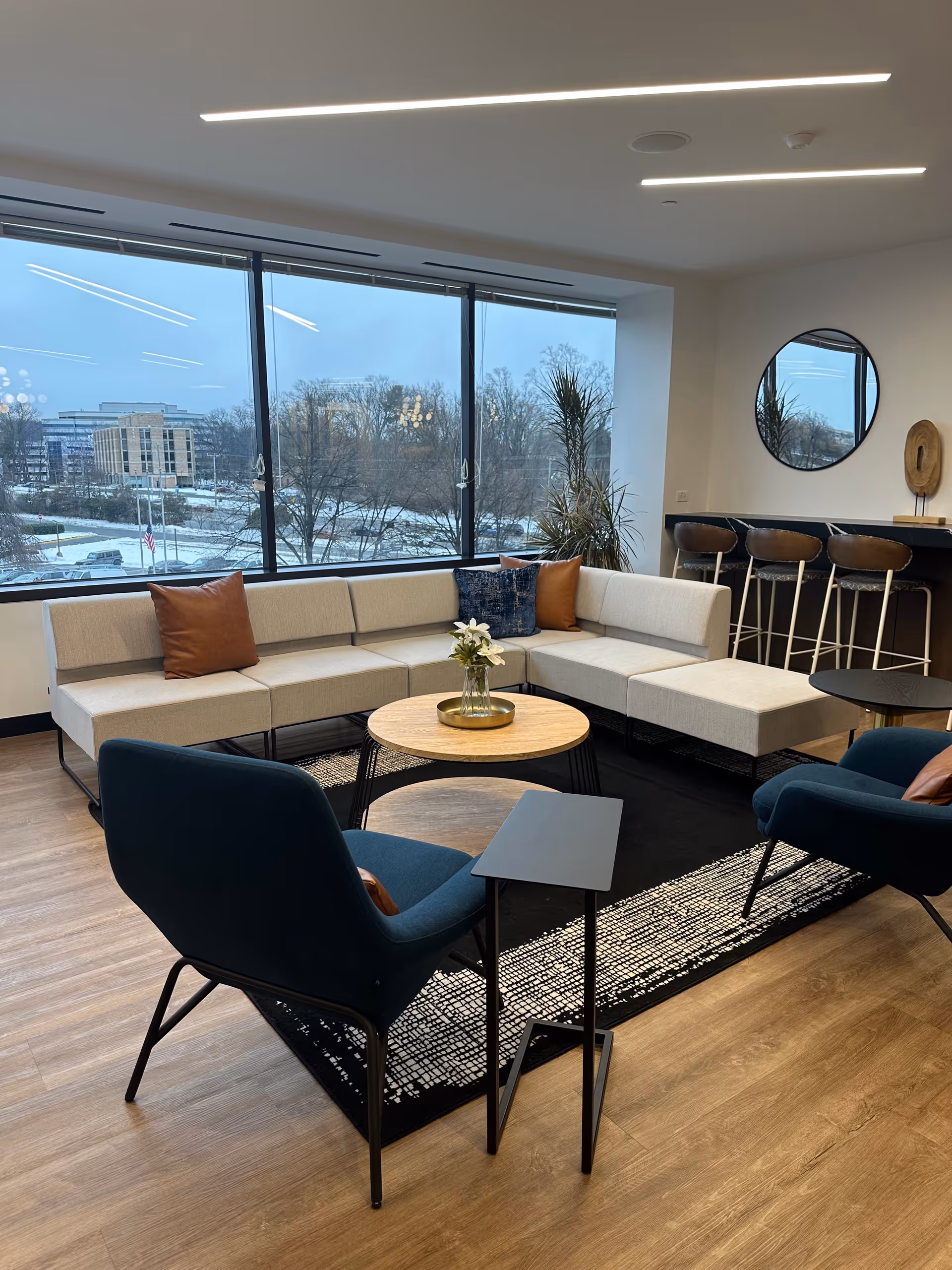 Bright, modern senior living lounge with beige sectional sofa, high chairs, and a large window view.