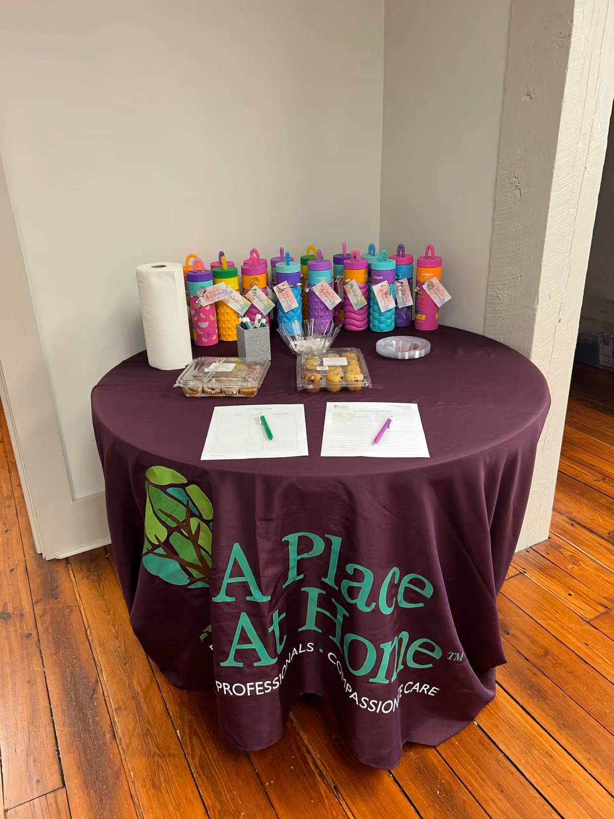 Round table with purple cloth, colorful water bottles, snacks, and sign-in sheets indoors.