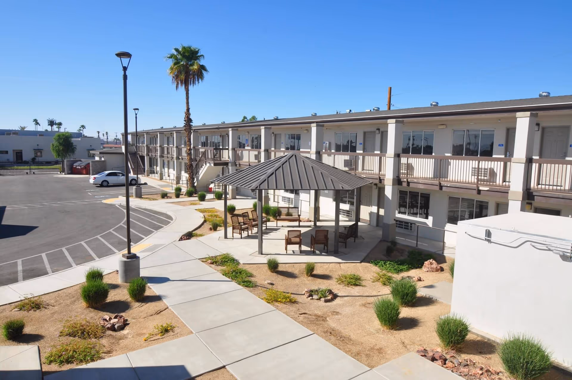 Courtyard of a two-story senior living building with a central gazebo, outdoor seating, walkways, parking area, and a tall palm tree.