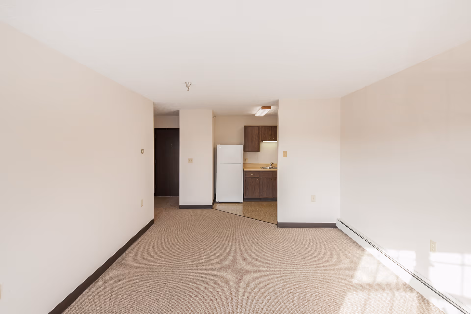 Empty room with beige carpet and walls, featuring a small kitchen area with a white refrigerator and dark wooden cabinets in the background. Sunlight is coming through a window on the right side, casting shadows on the wall and floor.