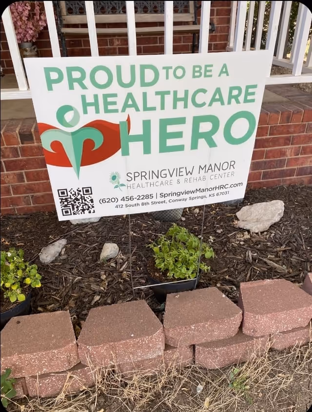 A yard sign that reads "Proud to be a Healthcare Hero" for Springview Manor Healthcare & Rehab Center placed in a flower bed in front of a brick porch.