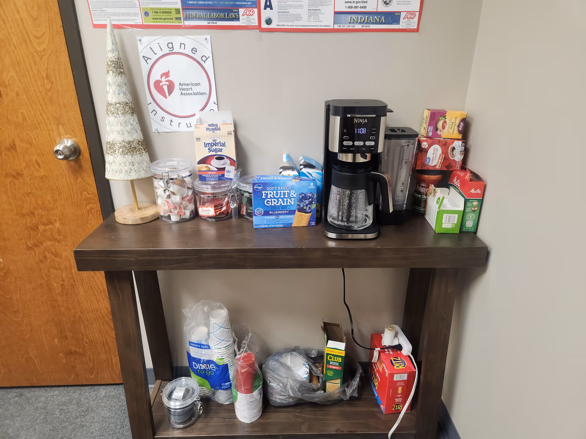 A dark wood table against a wall with a coffee maker, cups, and snack boxes.