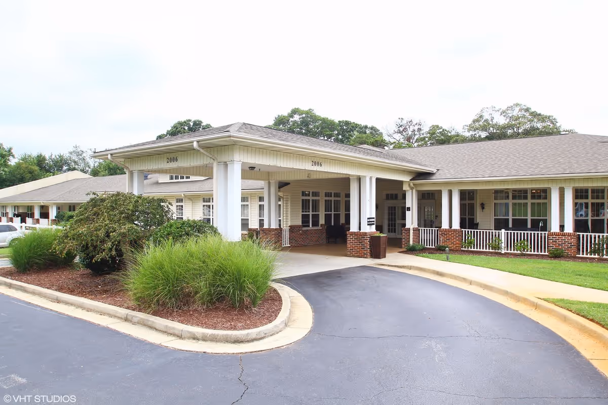 Exterior view of Pine Grove Senior Living facility showing a covered entrance with white columns and brick accents, surrounded by landscaped greenery and a curved driveway.