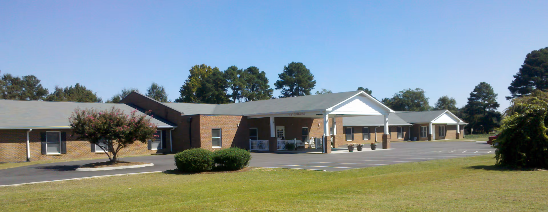 Exterior view of a single-story brick building with a covered entrance and multiple windows, surrounded by a parking lot, green lawn, trees, and clear blue sky.