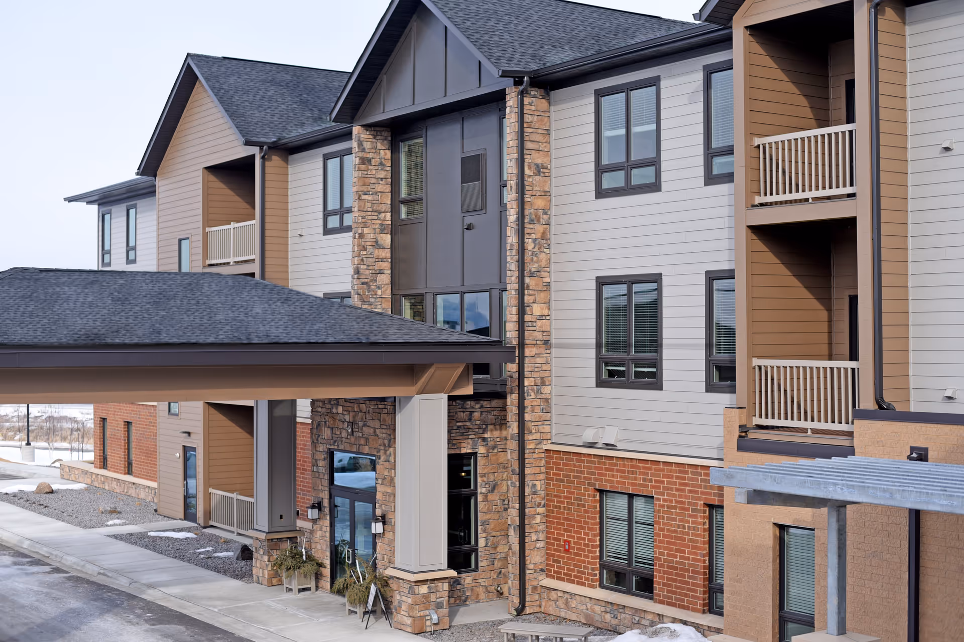 Exterior view of a modern senior living facility building with a covered entrance, multiple windows, balconies, and a combination of stone, brick, and siding on the facade. There is a sidewalk and some snow on the ground.