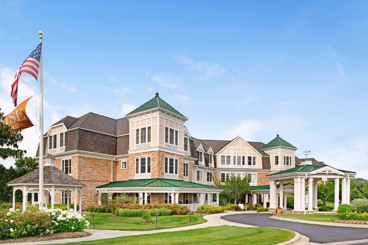 Exterior view of a senior living facility with a large multi-story building featuring stone and beige siding, green roofs, and multiple windows. There is a covered entrance with white columns and a driveway, surrounded by well-maintained green lawns, bushes, and flowers. An American flag and an orange flag are flying on a flagpole near the building.