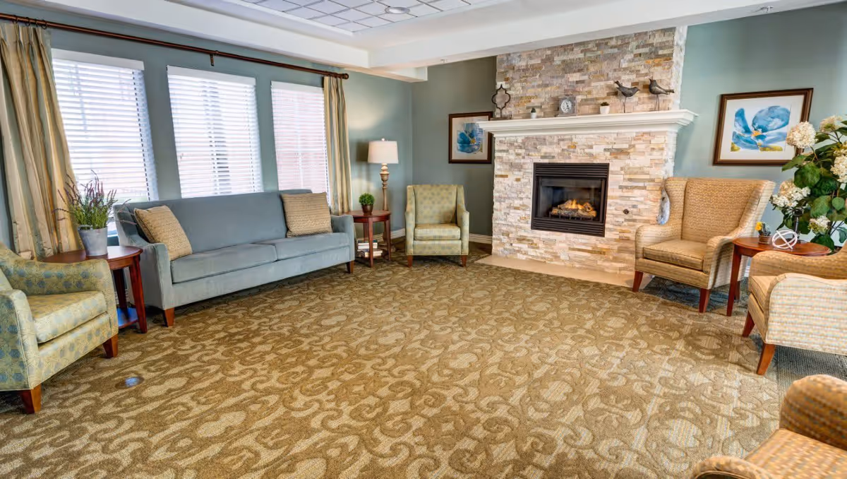 A cozy living room with a patterned carpet, a stone fireplace with a small clock and decorative birds on the mantle, a gray sofa with two cushions, four armchairs in different patterns, two side tables with lamps and plants, and three windows with blinds and curtains.