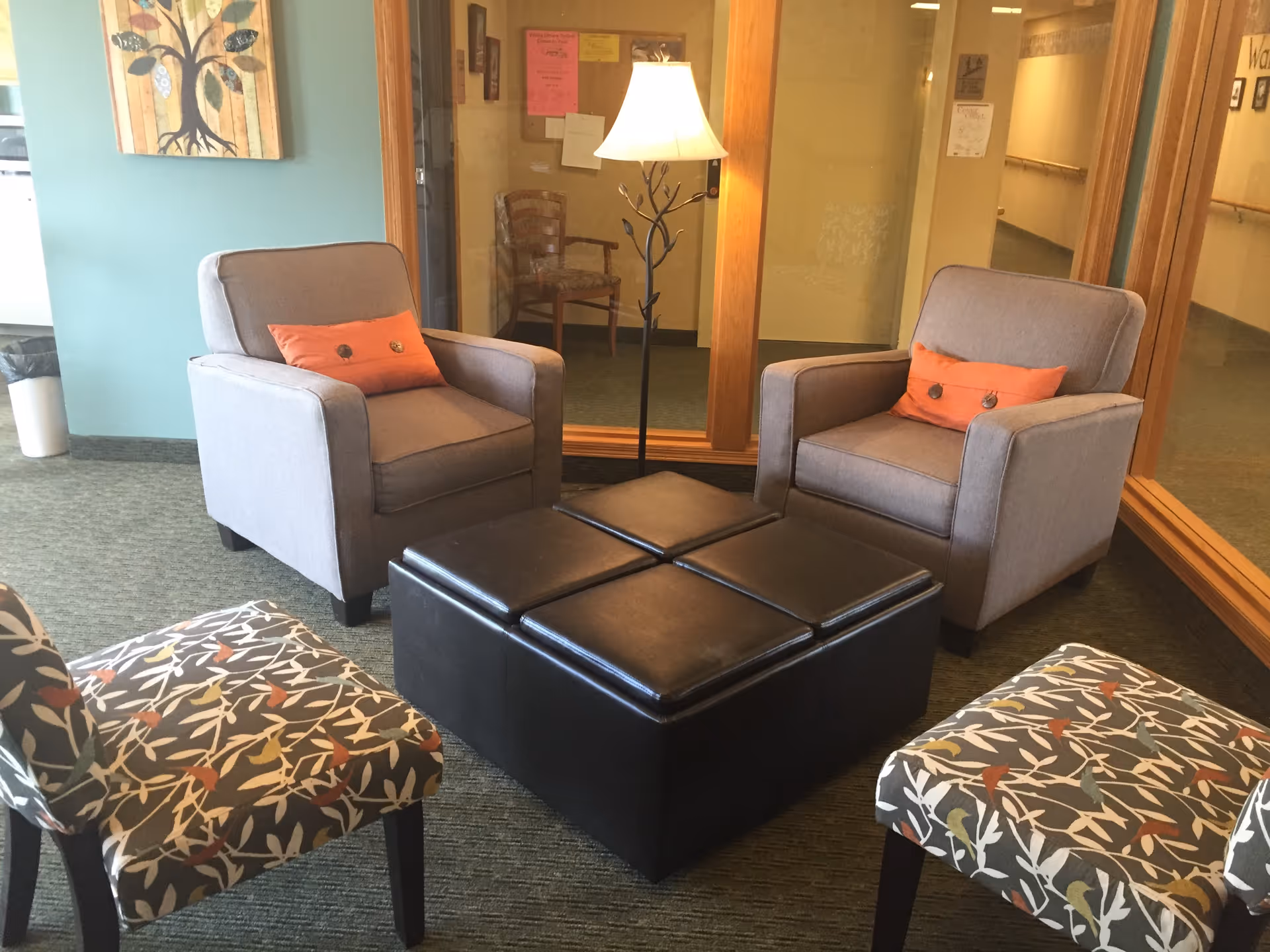 Cozy lounge area with two gray armchairs with orange pillows, two patterned side chairs, a square black ottoman, and a floor lamp.