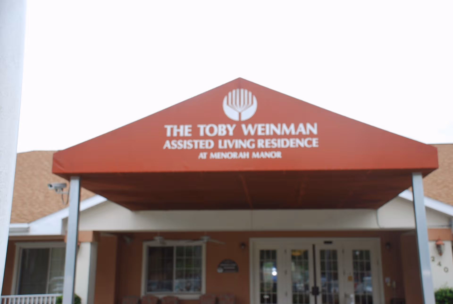 Red triangular canopy over the entrance reads The Toby Weinman Assisted Living Residence at Menorah Manor.